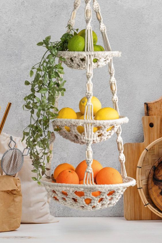 Hanging macrame fruit basket with oranges, lemons, limes, and greenery, kitchen decor with cutting boards and utensils.