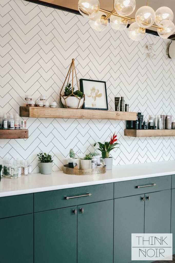 Stylish modern bathroom with dark green cabinets, wooden shelves, potted plants, and decorative items, herringbone tiles.