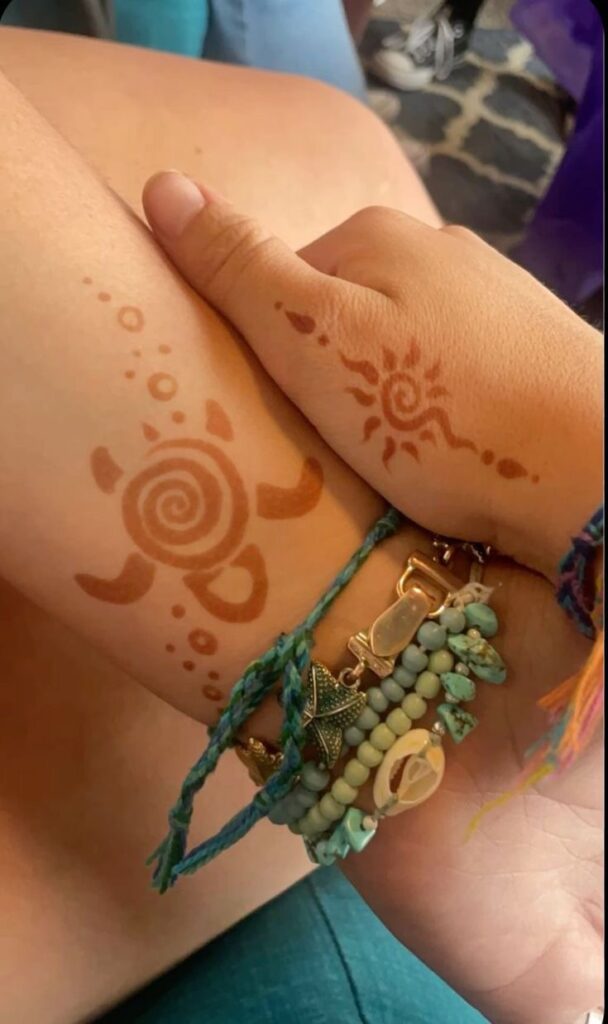 Hands with henna tattoos of a turtle and sun, wearing boho-style bracelets with beads, shells, and colorful threads.