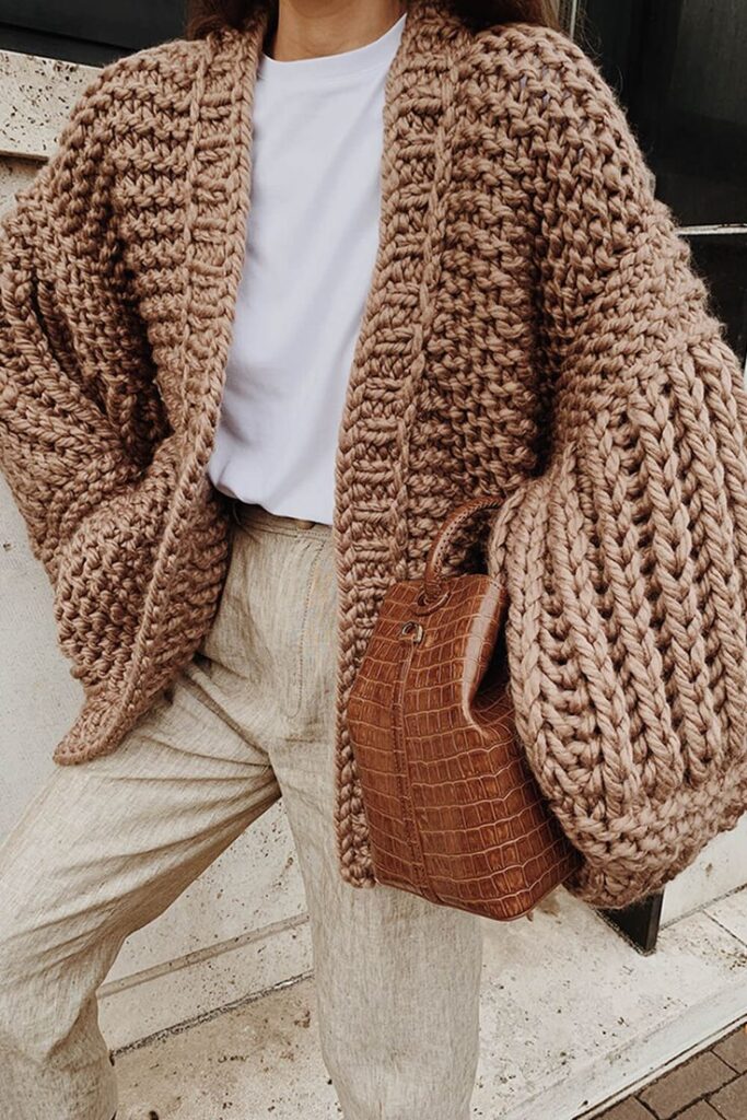 Woman wearing chunky knit cardigan, white t-shirt, beige pants, and brown leather handbag. Cozy autumn fashion outfit.