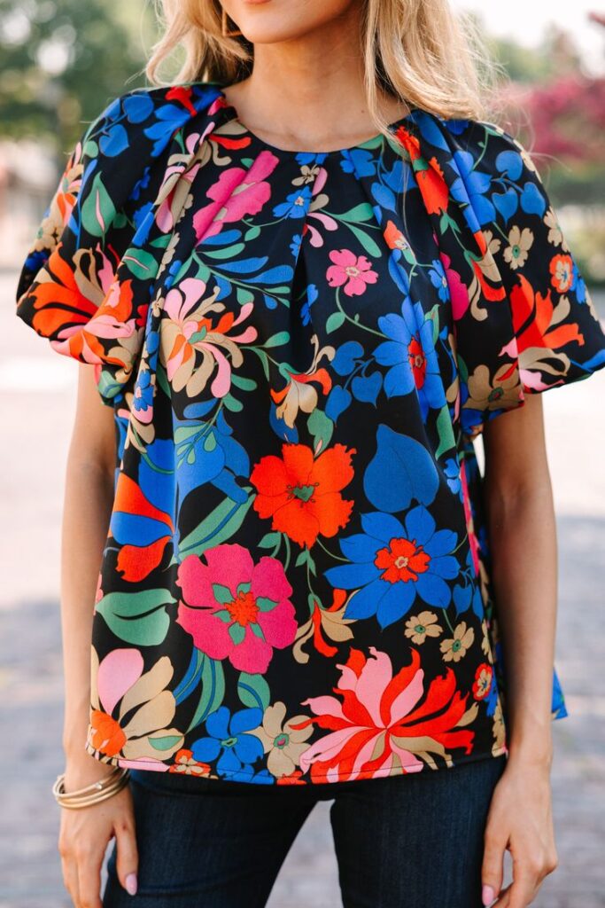 Woman wearing a vibrant floral print blouse with blue, red, and pink flowers on a black background, paired with dark jeans.