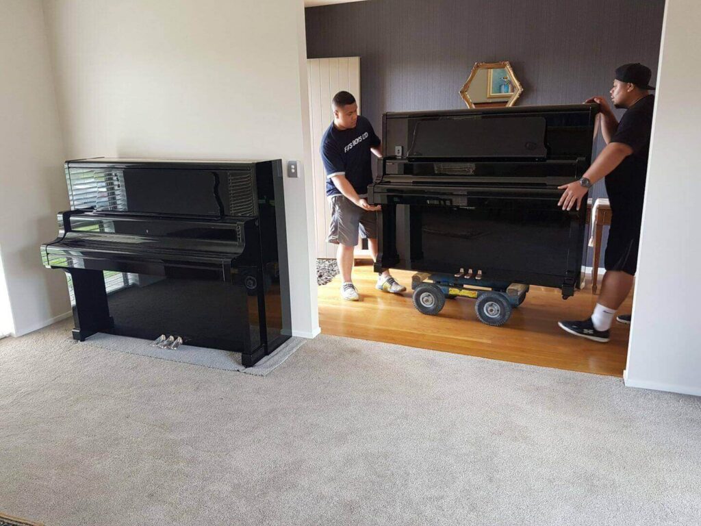 Movers carefully transporting a black piano on a dolly into a carpeted room. Another black piano is already in the room.
