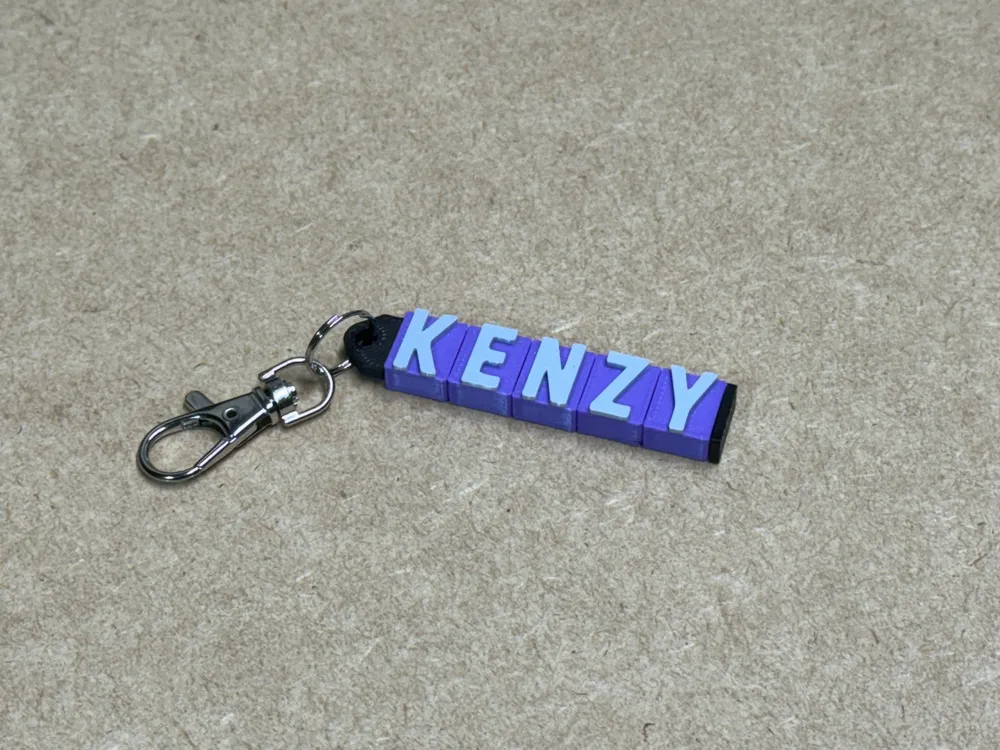 Purple keychain with the name KENZY in white letters, attached to a metal clasp, lying on a beige surface.