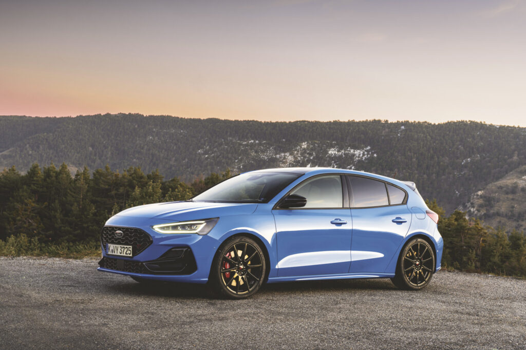 Blue compact car parked with mountain forest background at sunset, showcasing sleek design and stylish features.