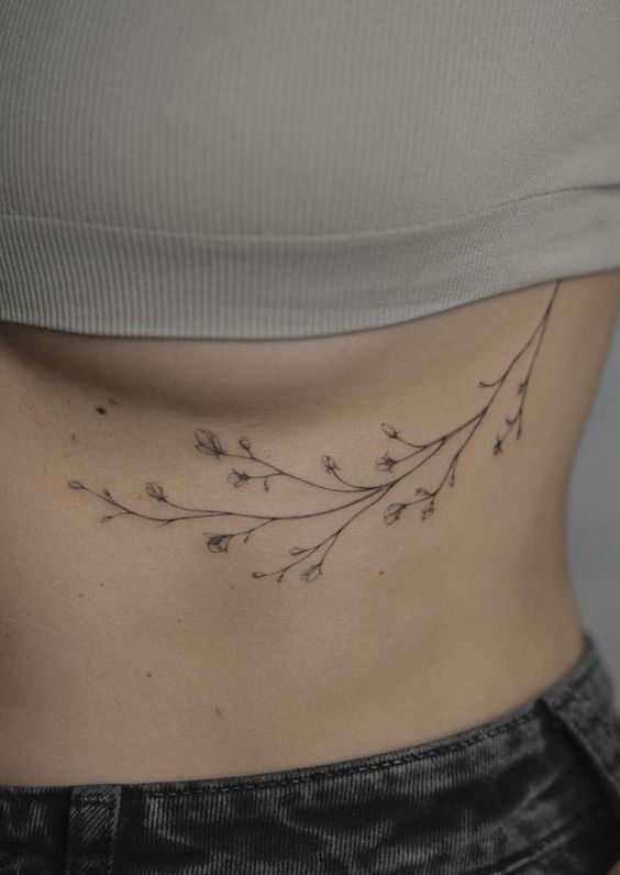 Minimalist floral branch tattoo inked on the side of woman's torso, under the ribcage, in black outline style.