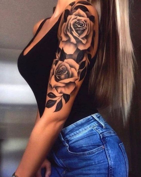 Woman with long hair and floral arm tattoo wears a black top and jeans, showcasing intricate rose and leaf design.