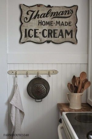 Vintage kitchen corner with sign reading Thalman's Home-Made Ice-Cream, cookware, wooden utensils, and towel hanging on hooks.