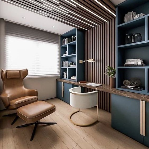 Modern home office with a leather chair, wooden desk, blue shelves, and stylish decor under natural light from a large window.