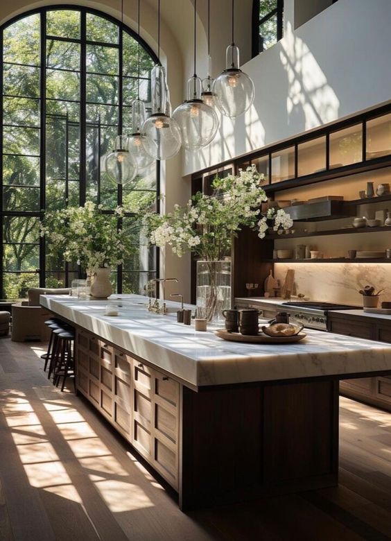 Modern kitchen with large windows, marble island, elegant lighting, and floral accents. Bright, airy, open-concept design.