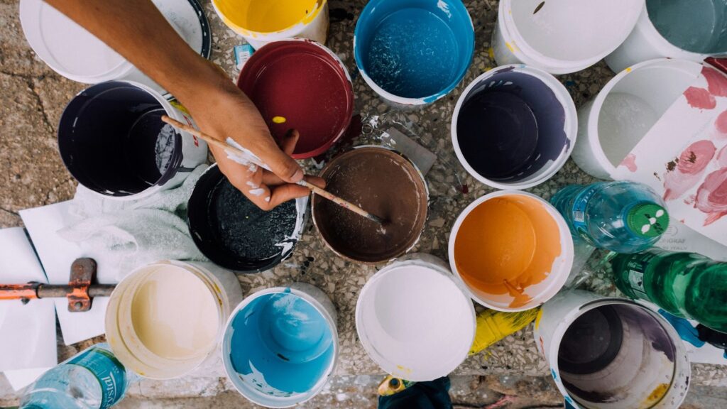 Various paint buckets in vibrant colors with a hand mixing brown paint using a brush. Ideal for art and creativity themes.