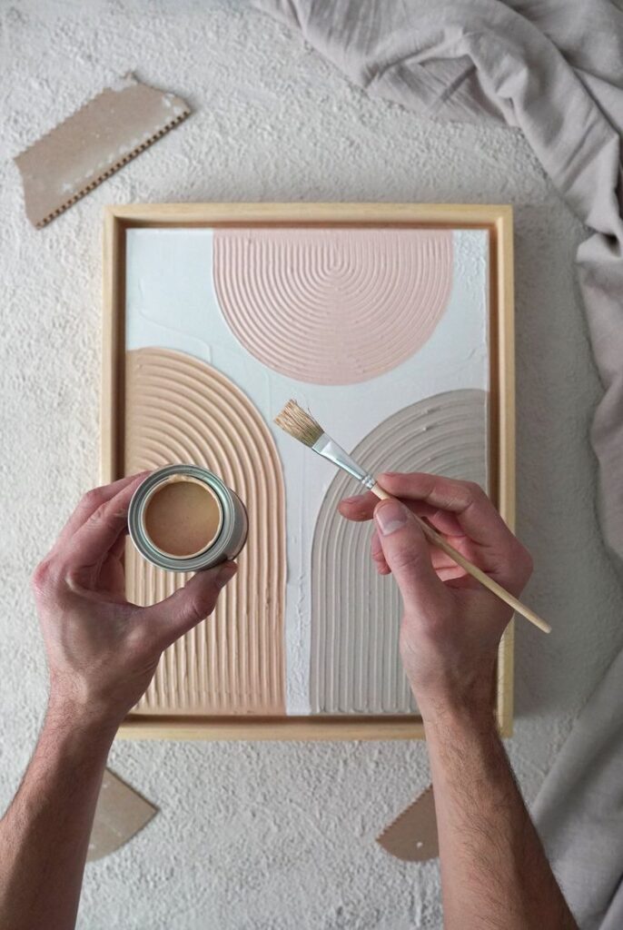 Hands holding a paintbrush and a paint cup creating textured artwork with pastel colors on a framed canvas.