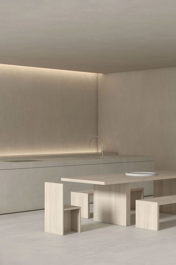 Minimalist kitchen with light wood dining table, matching bench, stools, modern faucet, and ambient lighting against beige walls.