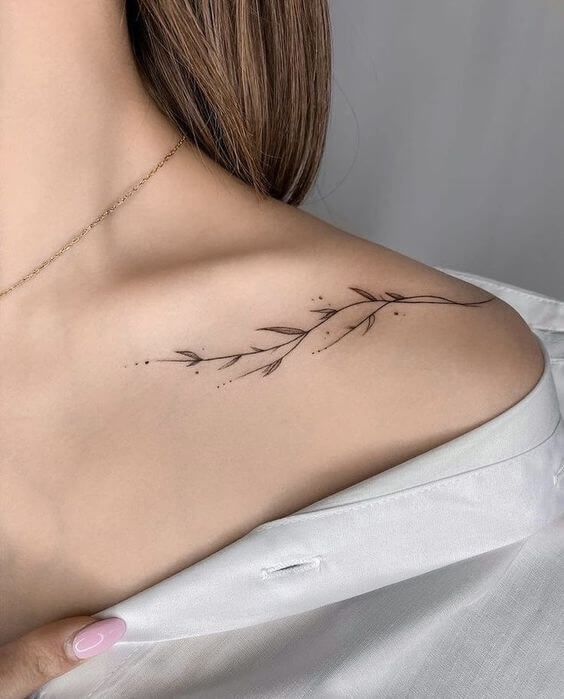 Delicate floral collarbone tattoo on a woman's shoulder, showcased in a minimalistic setting with a white shirt.