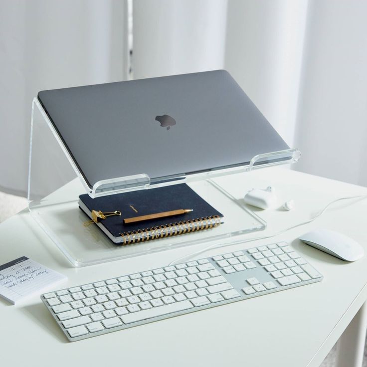 Ergonomic workspace with a laptop on a stand, keyboard, mouse, notebook, pen, and earbuds on a white desk.