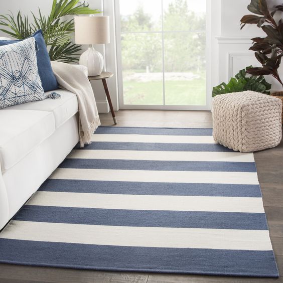 Modern living room with white sofa, blue and white striped rug, knitted pouf, and stylish decor near a large window.