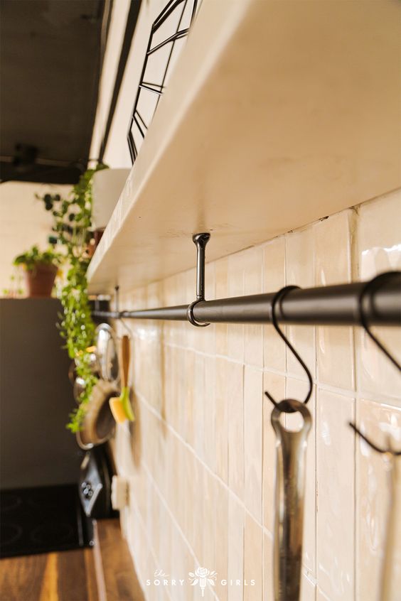 Modern kitchen with white ceramic tiles, black rail hooks, and potted plants on shelves, featuring utensils and a cozy atmosphere.