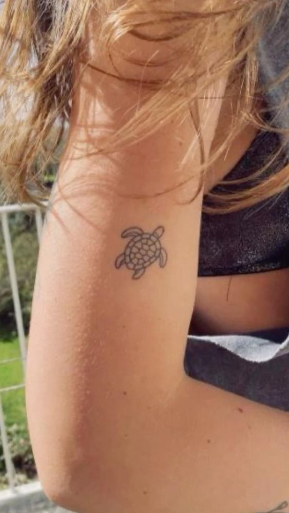 Close-up of a small turtle tattoo on a person's upper arm, with sunlight illuminating the tattoo and hair blowing in the breeze.