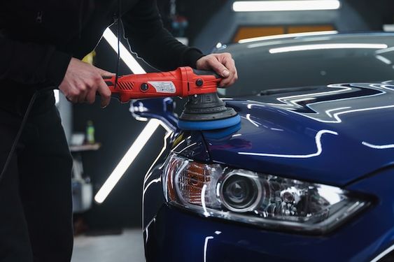 Person using a polisher to detail a shiny blue car in a garage with professional lighting.