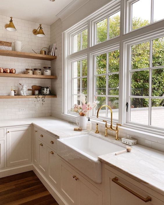 Bright farmhouse kitchen with white cabinets, open wooden shelves, gold fixtures, and a large window with garden view.
