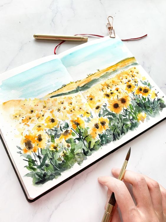 Watercolor painting of vibrant sunflowers in an art journal, with a brush and pen on a white table.