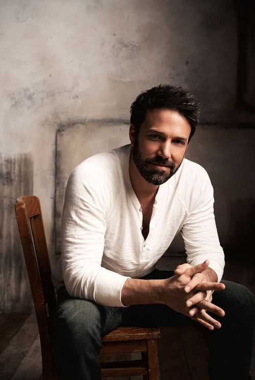 Man with beard and white shirt sitting on wooden chair against a textured background, looking at the camera.