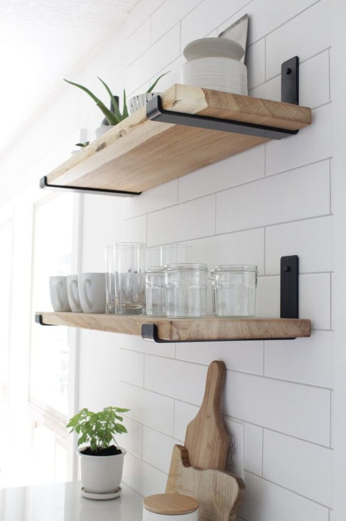 Minimalist kitchen with floating wooden shelves, glass jars, mugs, potted plant, and cutting boards on white subway tile wall.