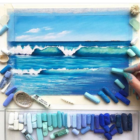 Artist creating a pastel drawing of ocean waves with various blue and turquoise pastel sticks scattered around.