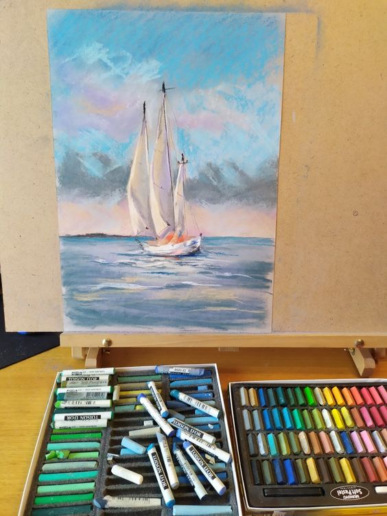 Pastel artwork of a sailboat on calm waters with colorful pastel chalks set on a table, showcasing artistic supplies.