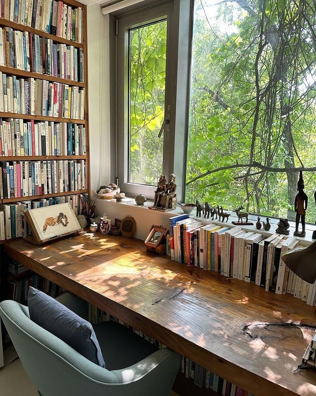 Cozy home office with a wooden desk, bookshelf, and a large window overlooking lush green trees, filled with sunlight.