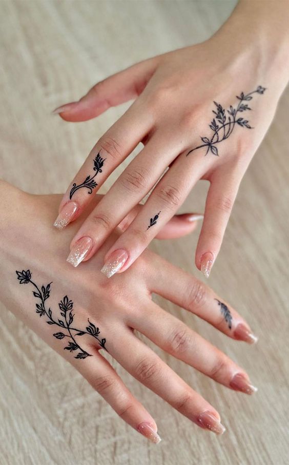 Hands with delicate floral tattoos and glittery nude nails on a wooden surface. Stylish nail art and henna design.