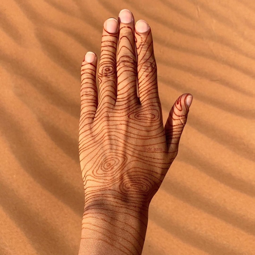 Hand with intricate henna design resembling wood grain patterns against a sandy desert background.