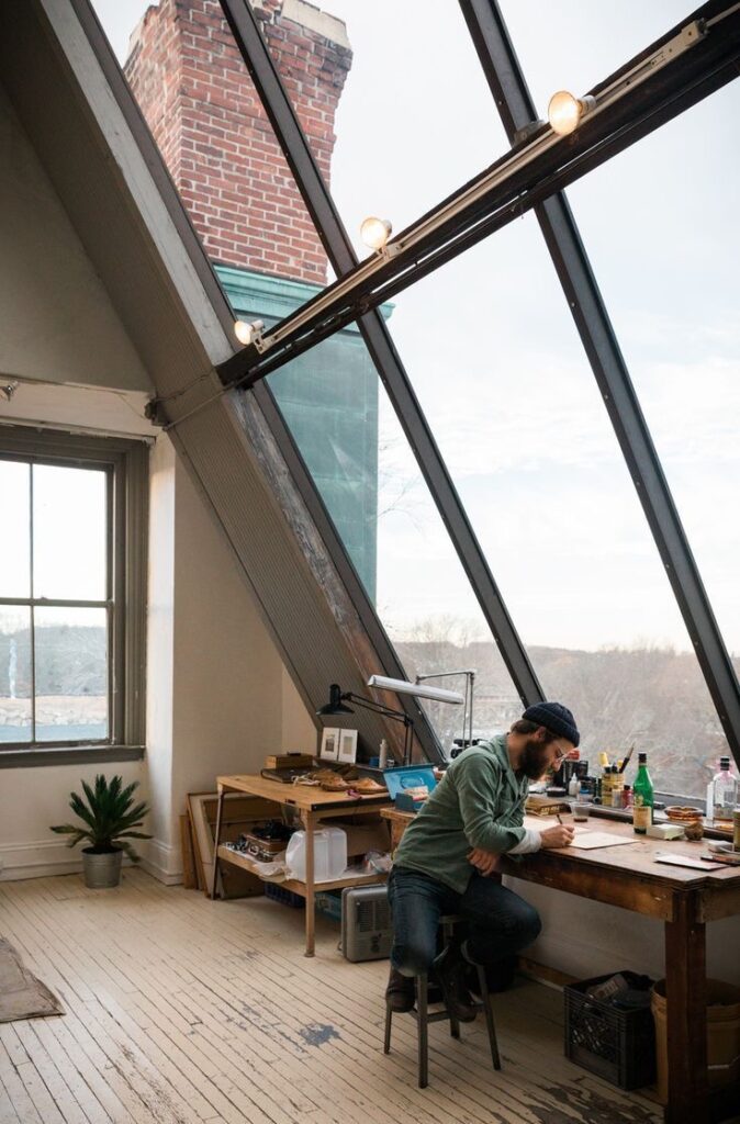 Artist working in sunlit studio with large windows, high ceiling, and rustic wooden furniture, surrounded by artistic materials.