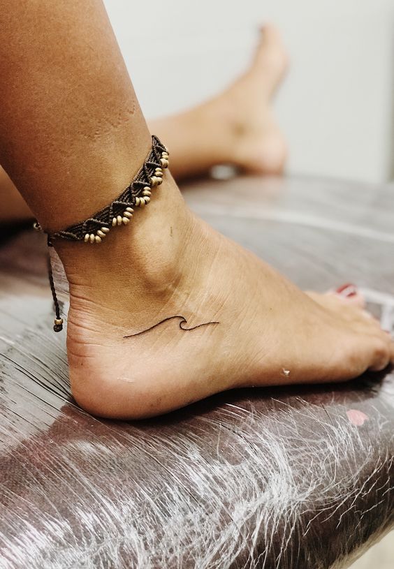 Ankle tattoo of a wave with beaded anklet bracelet on a person’s foot, highlighting minimalist surf-inspired design.