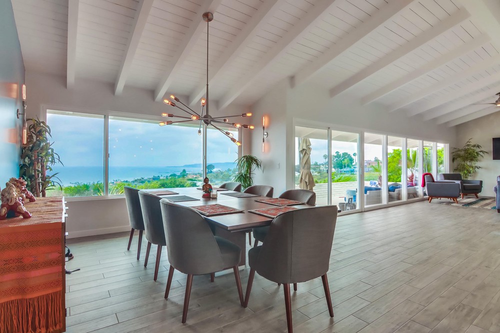 Stylish dining room with ocean view, modern furniture, large windows, vaulted ceiling, and contemporary chandelier.