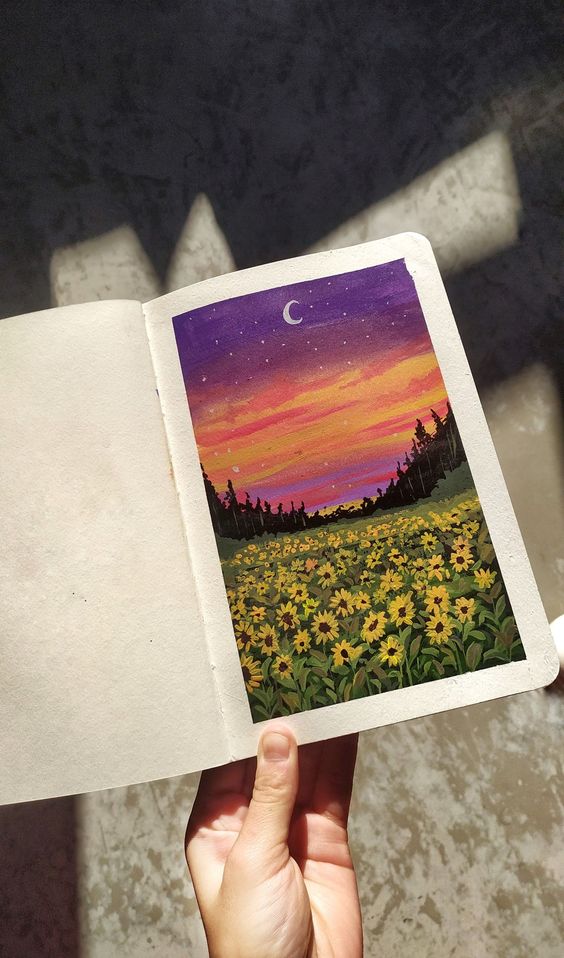 Hand holding an open sketchbook with colorful painting of a sunset over a field of yellow flowers and crescent moon.