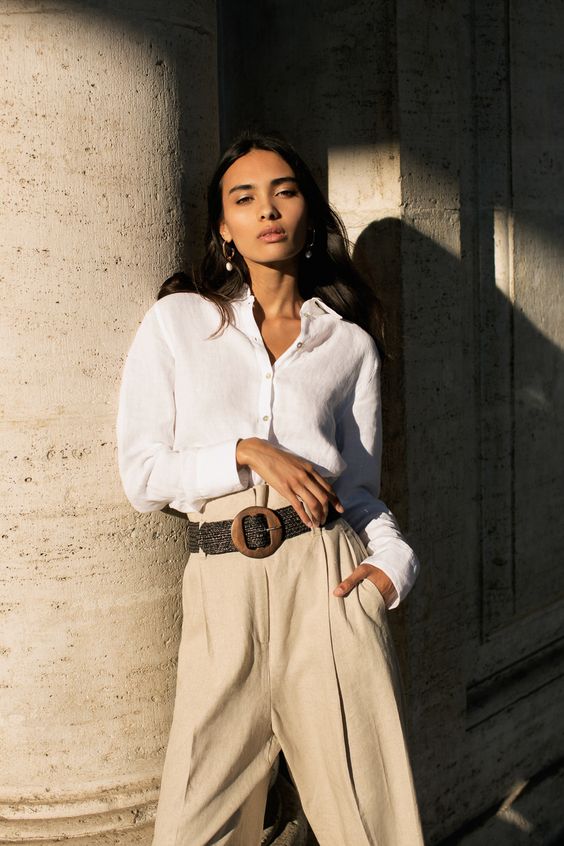 Woman in white shirt and beige pants, leaning on stone pillar with sunlight casting shadows. Fashionable and elegant look.