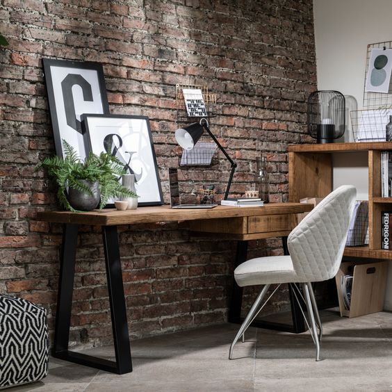 Modern home office with a rustic wooden desk, white quilted chair, and decor against an exposed brick wall. Industrial chic style.