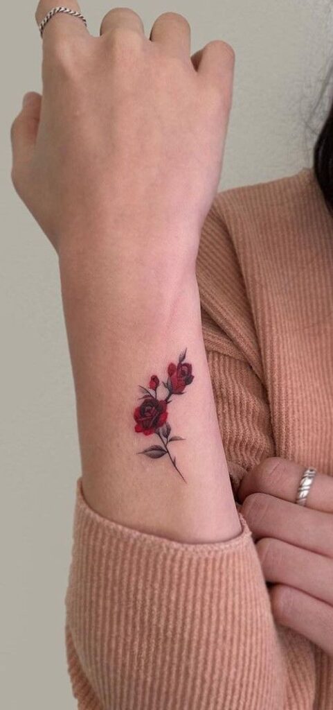 Minimalist red rose tattoo on the inner wrist of a person wearing a beige ribbed sweater and silver rings.