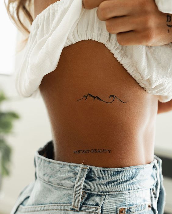 Minimalist side rib tattoos featuring a mountain wave design and FANTASY = REALITY text, worn by a person in denim shorts.