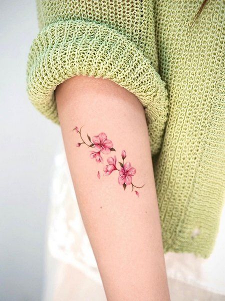 Close-up of a delicate pink flower tattoo on an arm, with the person wearing a light green knitted sweater.