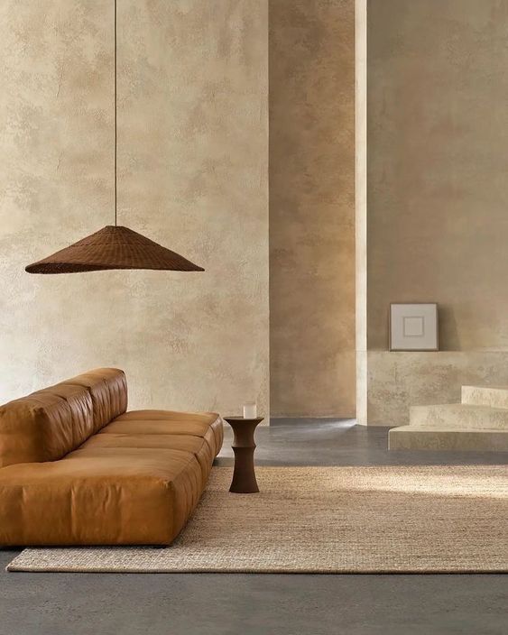 Minimalist living room with brown leather sofa, textured beige walls, pendant light, and woven rug, exuding warm, natural tones.