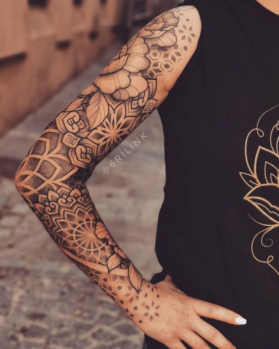 Arm with intricate floral and geometric sleeve tattoo in black ink, woman wearing black tank top with subtle design.