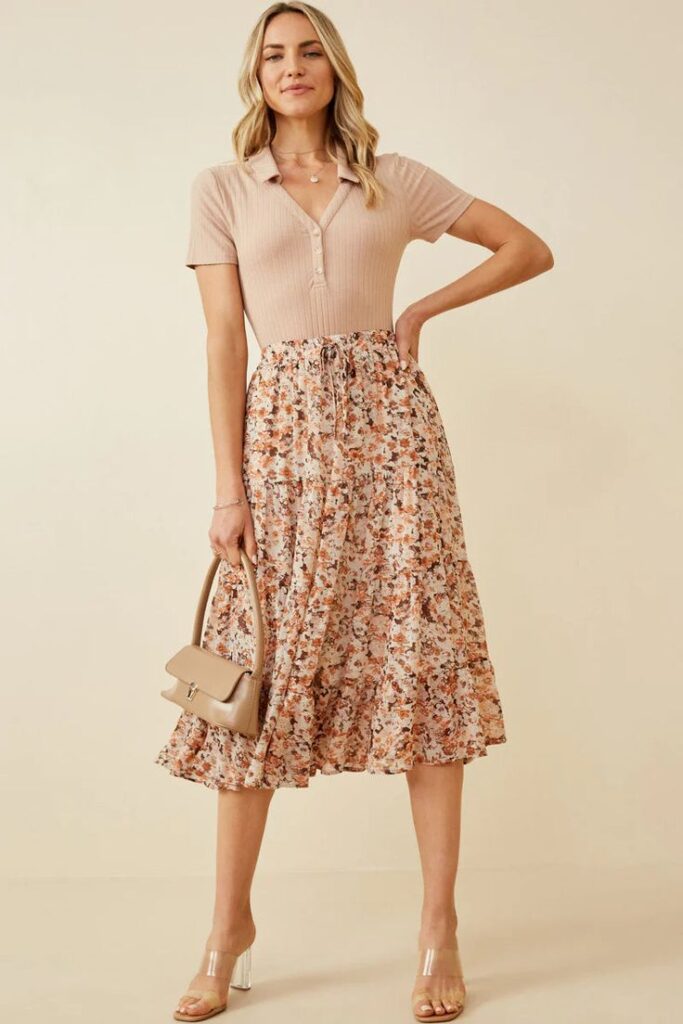 Woman in a beige top and floral skirt holding a beige handbag, posing in a neutral background, fashion outfit.