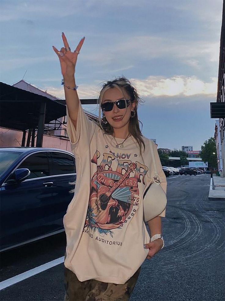 Woman in graphic tee making rock gesture, wearing sunglasses, with car and urban background, during a cool evening.