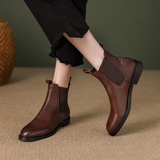 Woman wearing stylish brown leather ankle boots with black pants on beige carpet background. Fashionable women's footwear.