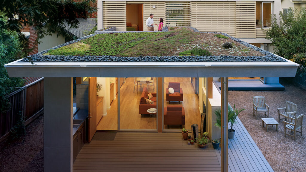 Modern house with a green roof and spacious living area, featuring a deck and outdoor seating, with two people on the balcony.