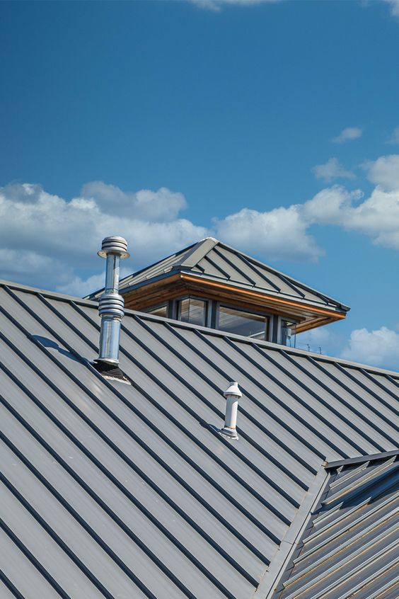 Modern metal roof with skylight and ventilation pipes under a bright blue sky with scattered clouds.