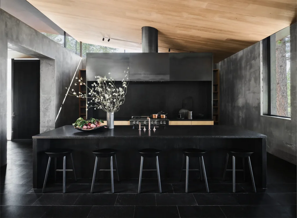 Modern black kitchen with large island, bar stools, elegant flower vase, and natural light from large windows.