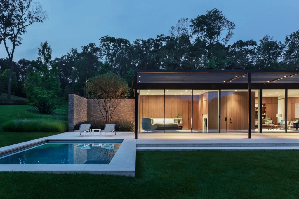 Modern luxury home with glass walls, wooden interior, and a poolside lounge area surrounded by lush greenery at dusk.