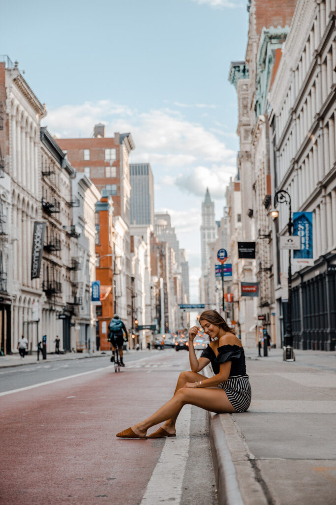 Young woman sitting on a curb in a bustling city street, with tall buildings and a cyclist in the background on a clear day.